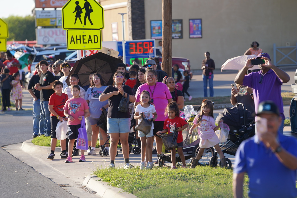 Many people wait in line on a sidewalk, with children holding hands and others holding umbrellas.