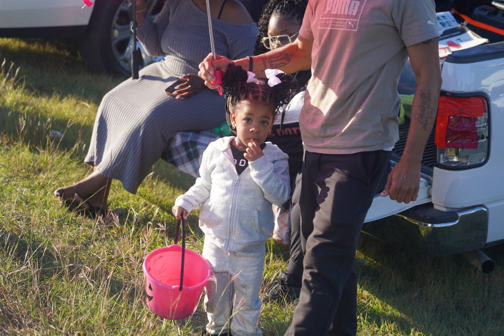 Two adults and a small child stand in grass. A girl holds a pink bucket. A vehicle is behind them.