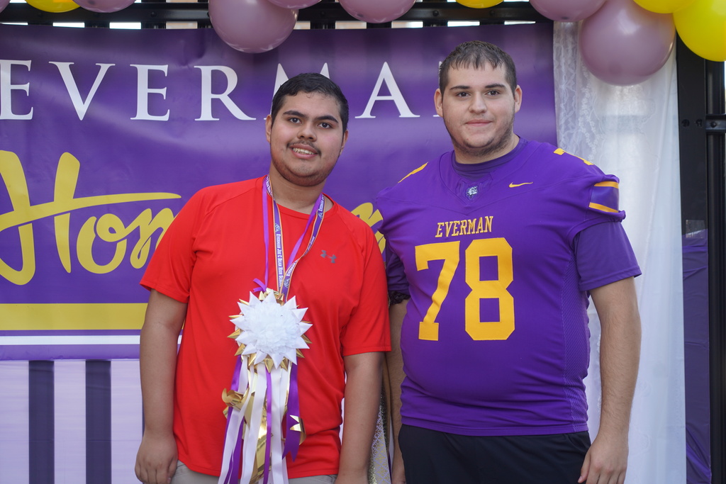 Two young men, one wearing a red shirt and the other a purple jersey with the number 78, stand before a banner with balloons.