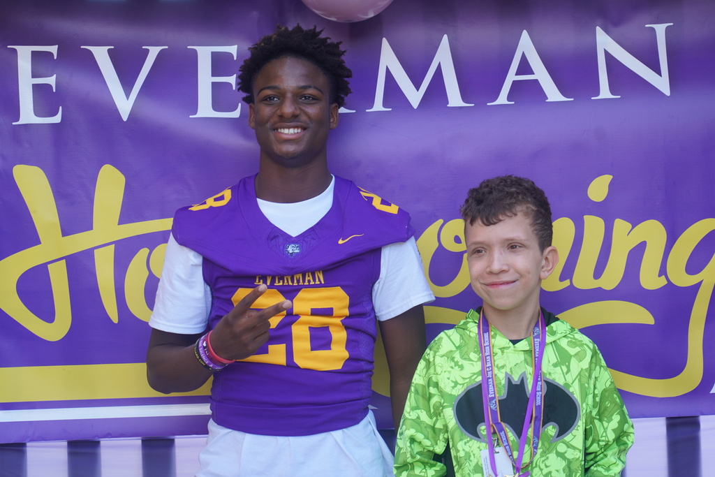 Two kids standing next to each other. One is wearing a football uniform and the other a superhero costume.