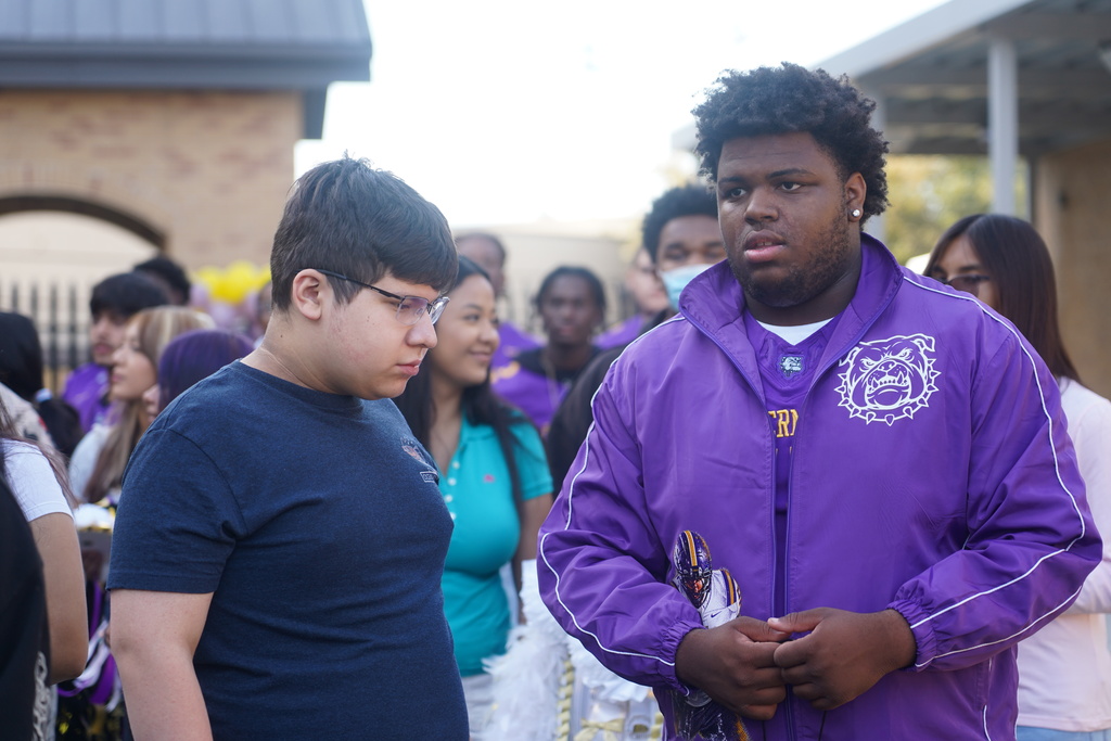 Two students stand in front of a building with others; one wears glasses, the other a purple jacket.