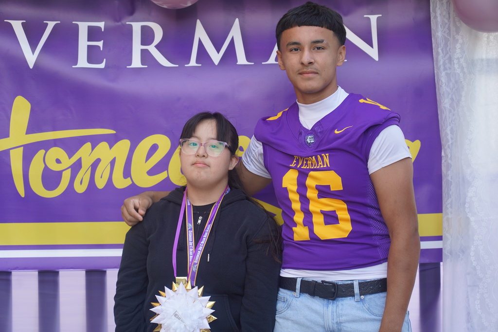 Two students stand in front of a purple and yellow banner, one wearing a football jersey with the number 16, the other with a necklace and medal.