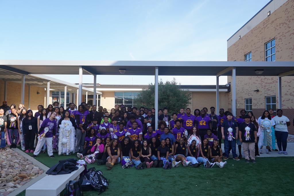 Group of people in purple and white shirts and white pants gathered on green artificial turf in front of a building.