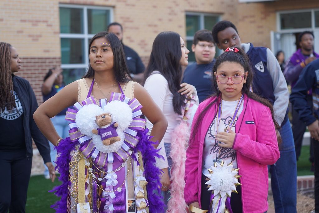 Several students stand together in front of a building, two wearing elaborate decorative costumes. One student has a stuffed animal in her costume.