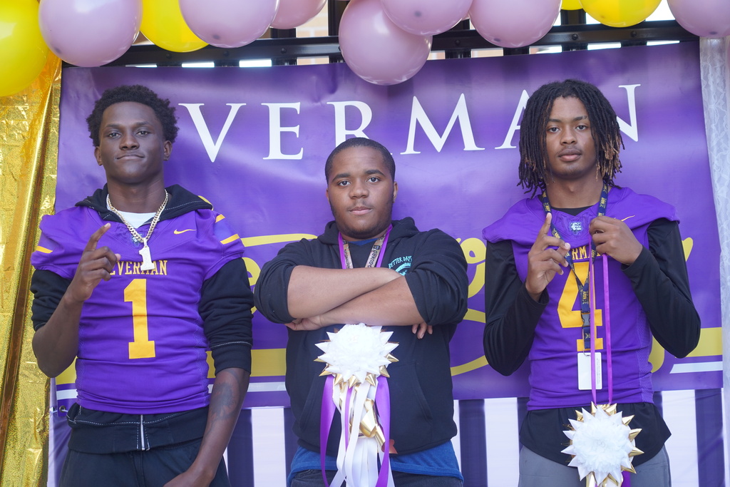 Three male athletes in purple jerseys stand under a banner reading "Verman". They hold medals and pose with arms crossed.