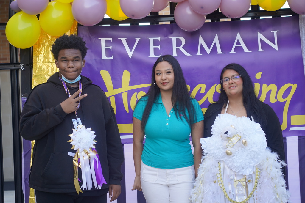 Three students pose in front of a purple banner with gold letters and yellow balloons.