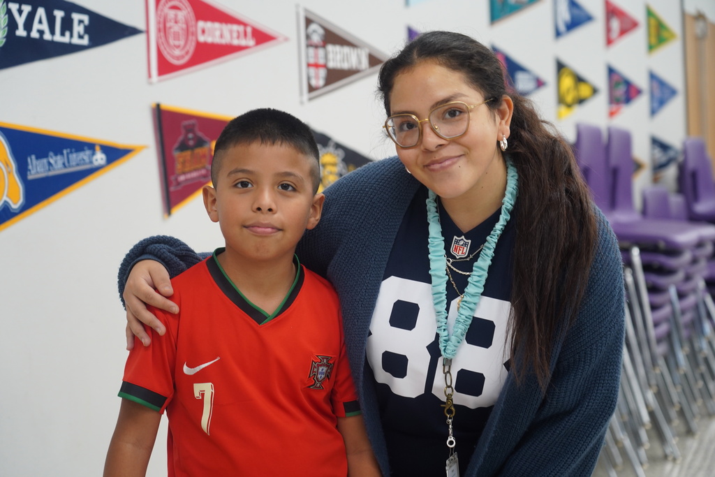 A woman and a boy in a classroom pose for a photo with university pennants in the background.