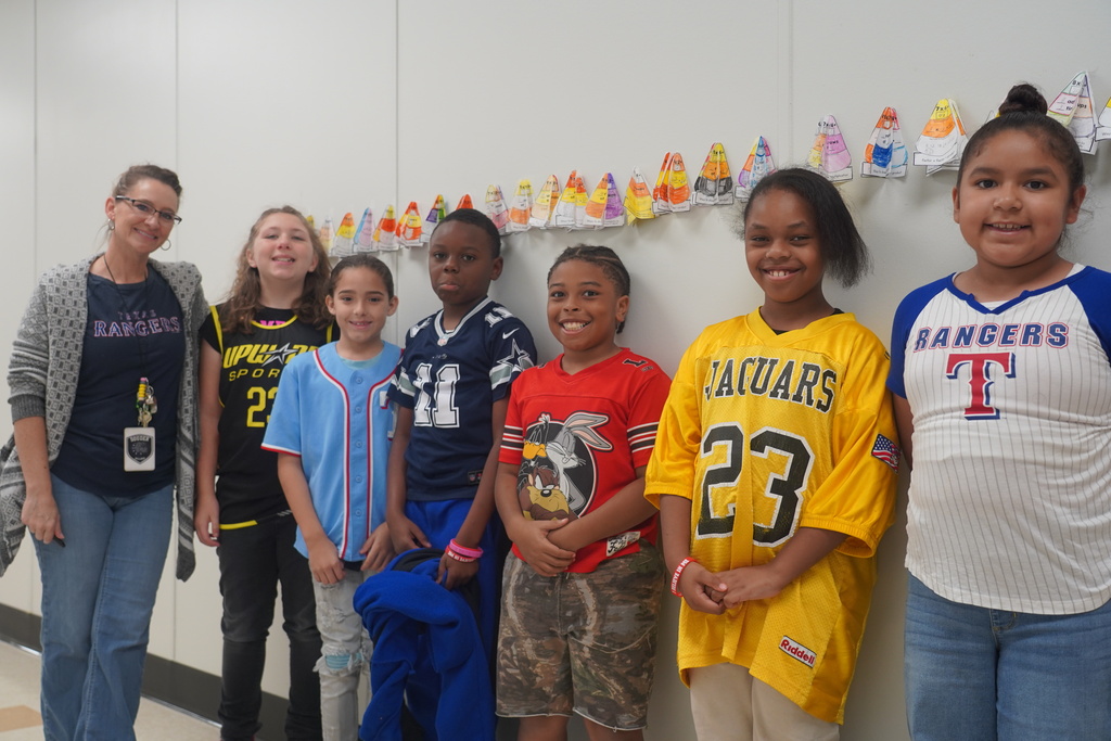 Students wearing jerseys with numbers and team names stand in a hallway next to a wall adorned with colorful Easter decorations.