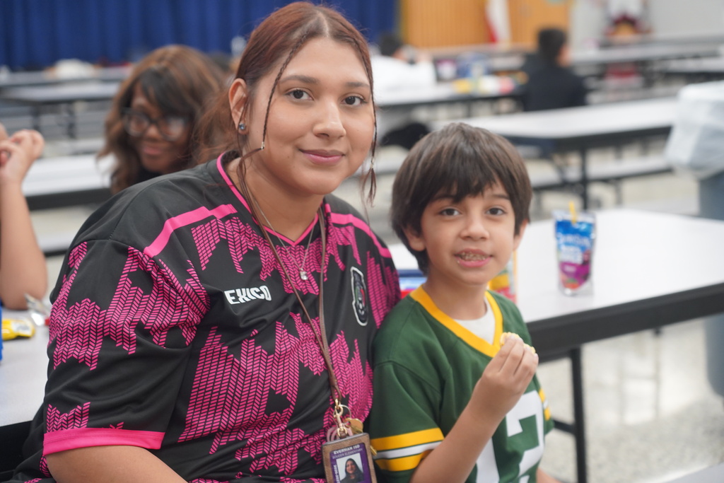 Woman and child sit at a table. Woman wears a black and pink shirt. Child wears a green shirt with yellow trim.