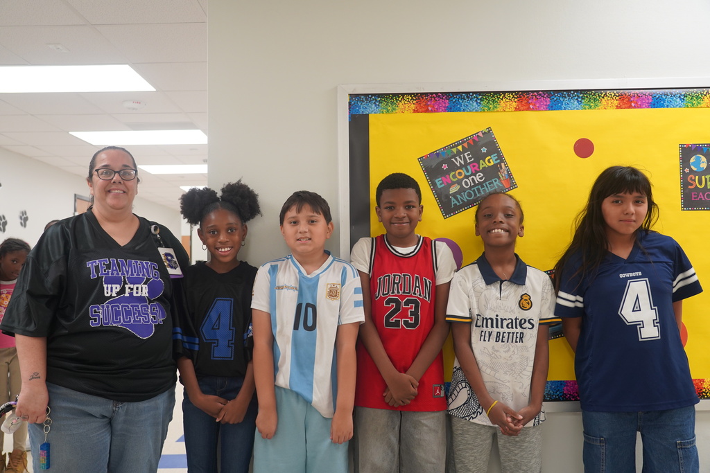 A woman and five children stand together. The wall behind them has a yellow bulletin board with multicolored papers.