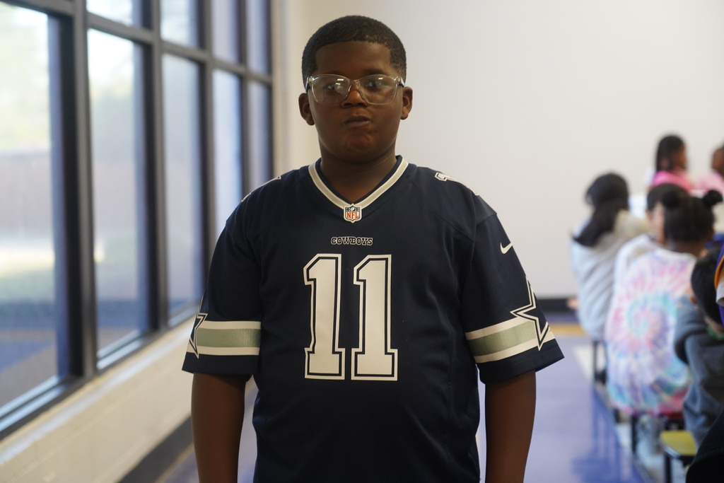 A student in a sports jersey stands in a hallway with others sitting on benches.