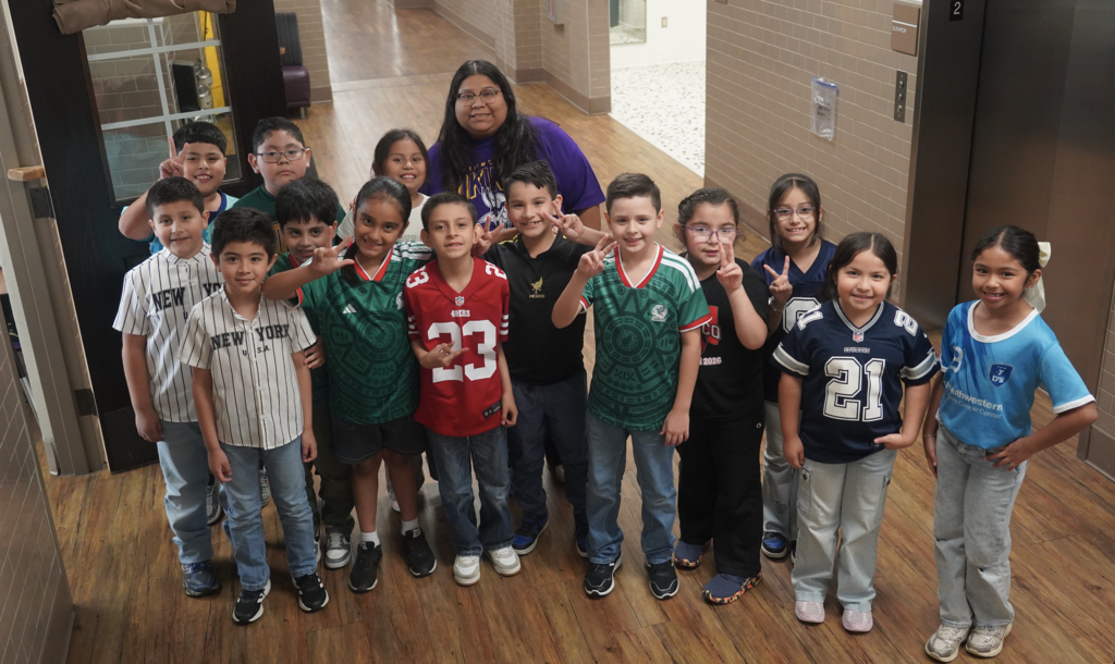 A group of students wearing various sports jerseys pose in a hallway with a teacher.