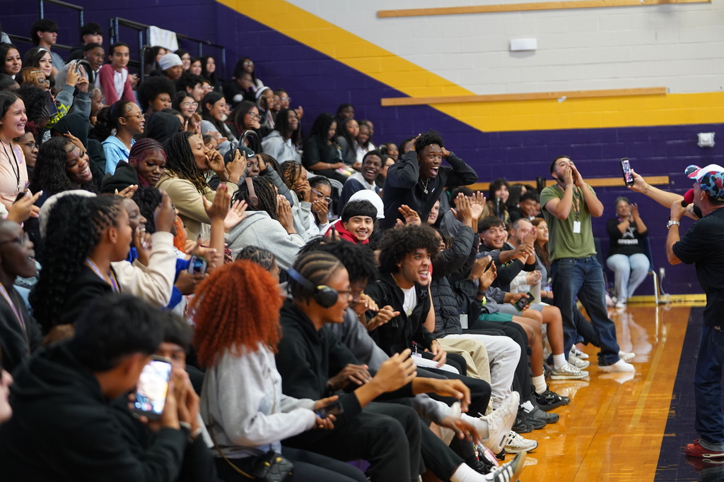 Audience seated in bleachers, some clapping, seated on basketball court with a speaker standing on the right.