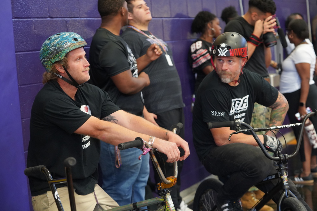 Two men wearing helmets and black t-shirts sit on bikes; a crowd of people stands behind them watching.