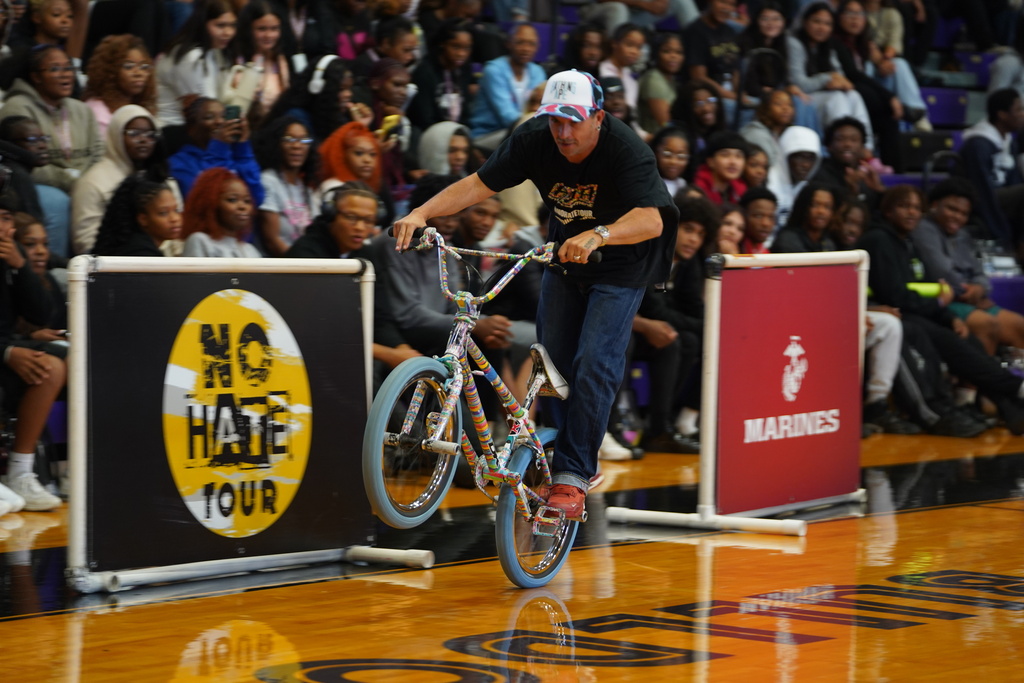 A man on a bicycle performs stunts in a gymnasium. People watch from the bleachers.