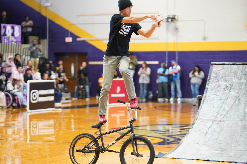 A person stands on a unicycle in the middle of a basketball court with onlookers behind him.