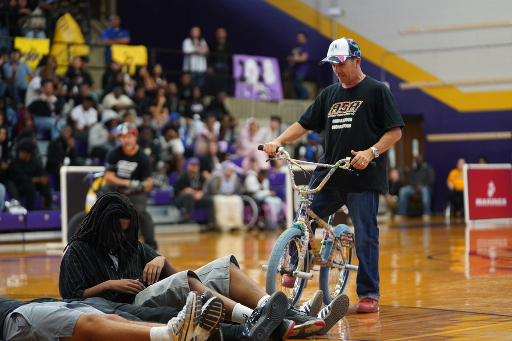 A man stands next to a bicycle while three people lie on the ground in front of a seated audience.