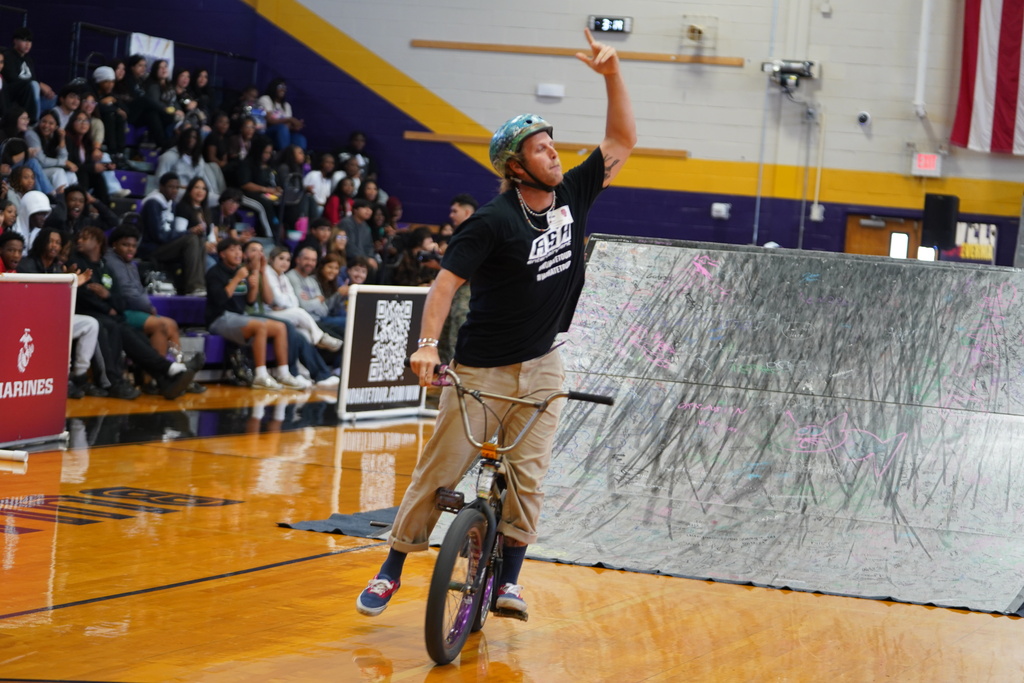 Man on bicycle riding on ramp in basketball court; crowd sitting on bleachers in background.