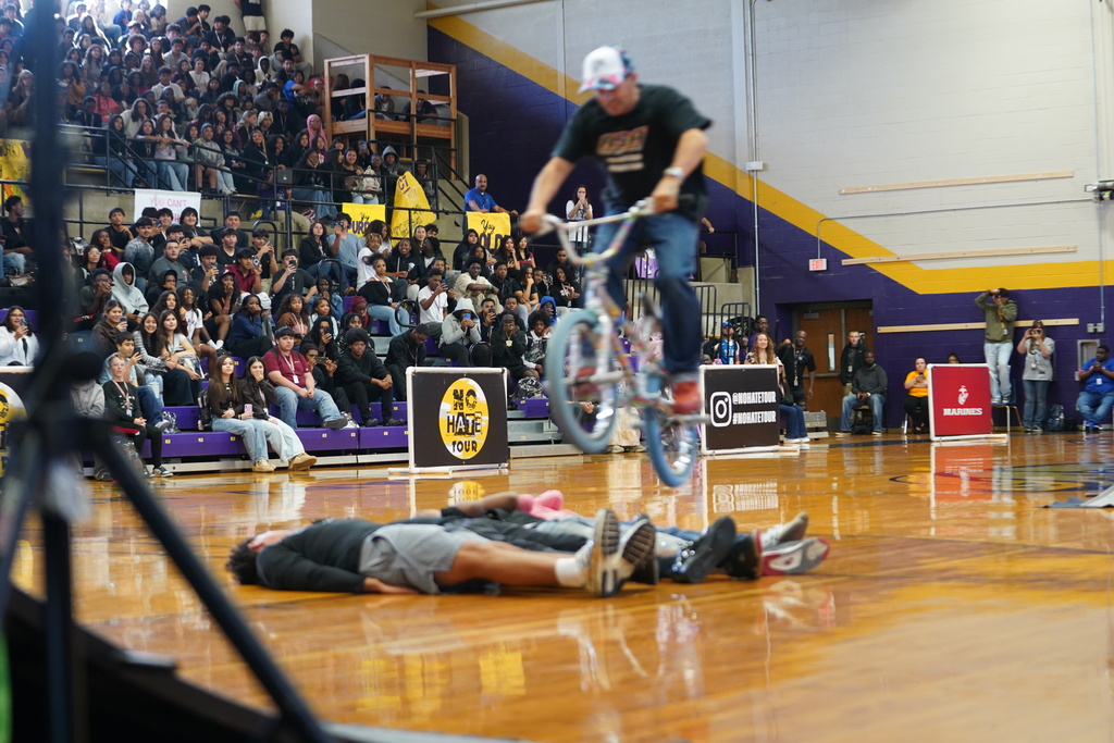 People in stands watching a man on a bicycle perform tricks over a person lying on the floor.
