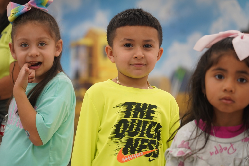 Three young children, each with different hair accessories, pose for the camera against a mural background.
