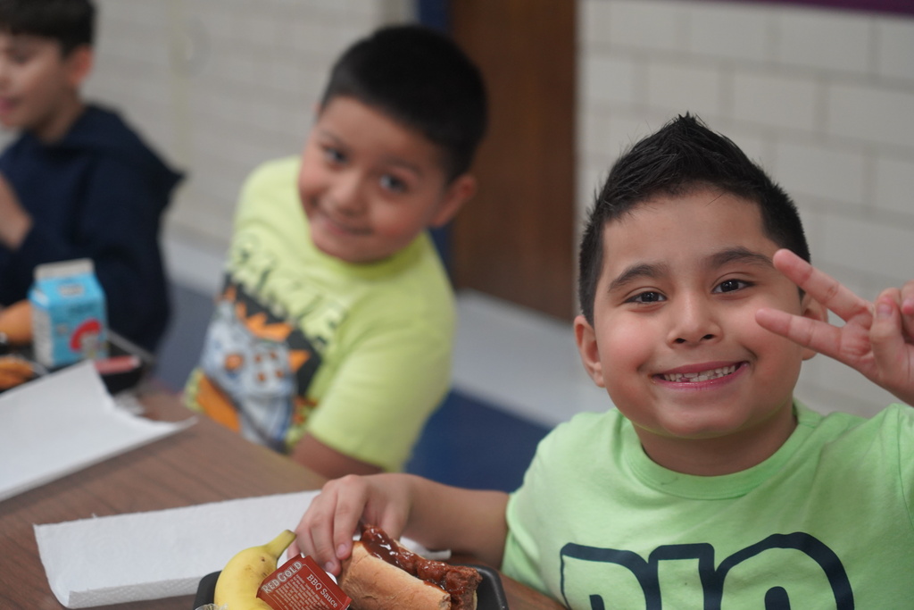 Two boys seated at a cafeteria table with food, one holding a banana and hot dog.