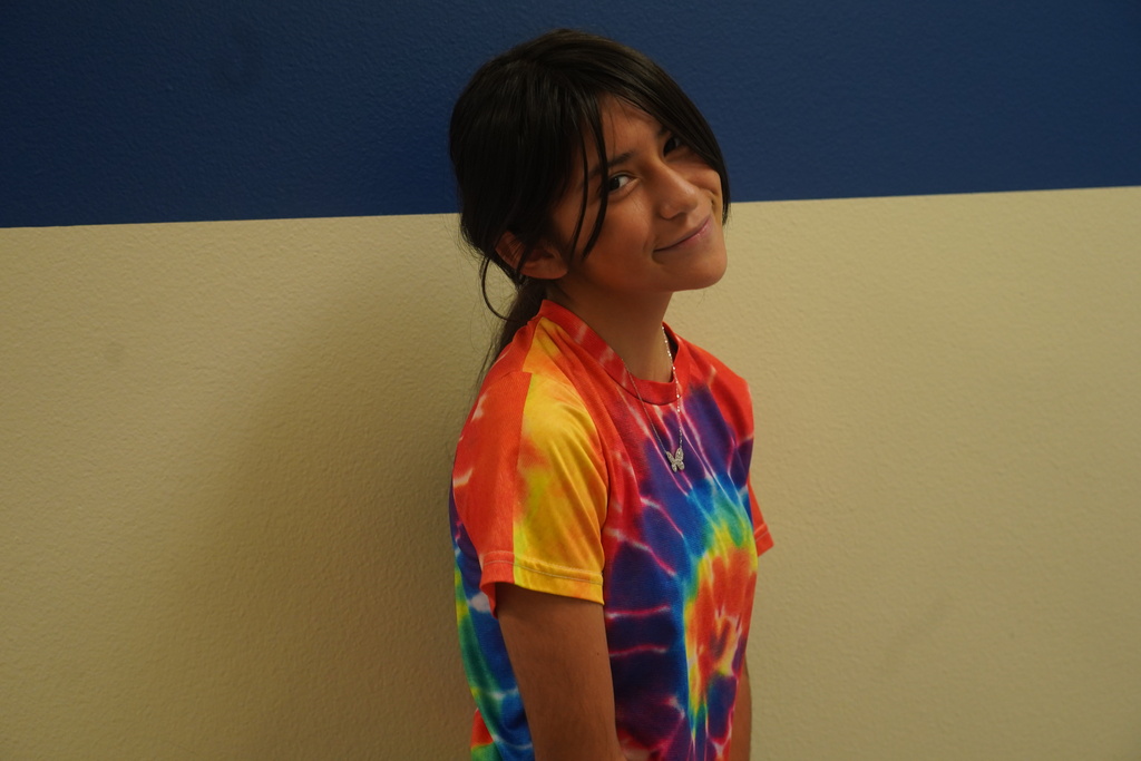 A student with short hair and a tie-dye shirt stands against a white and blue wall.