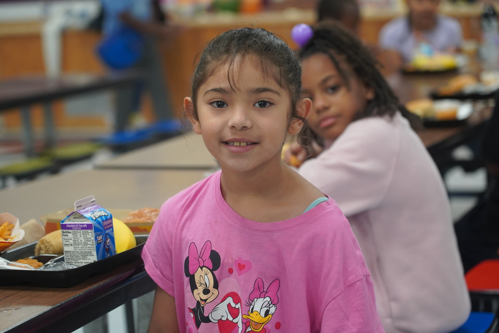 Girl in a pink shirt with Minnie Mouse stands in cafeteria. Another child sits behind her, eating from a tray.