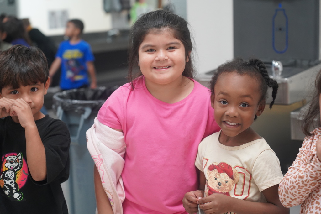 Four children smiling, two girls and two boys, in a hallway. One girl wears a pink shirt, the other a white shirt.