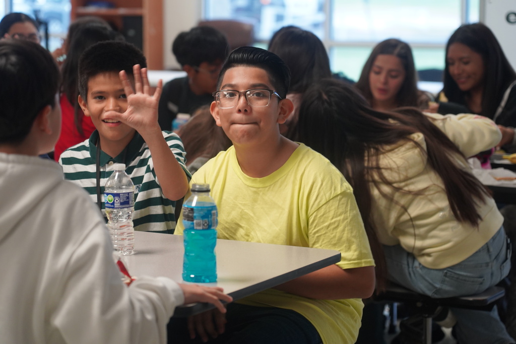 Children sit at tables in a classroom. A boy waves. A table has two water bottles.