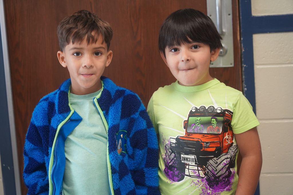 Two boys in front of a brown door; one wears a blue jacket, the other a green shirt with a vehicle graphic.