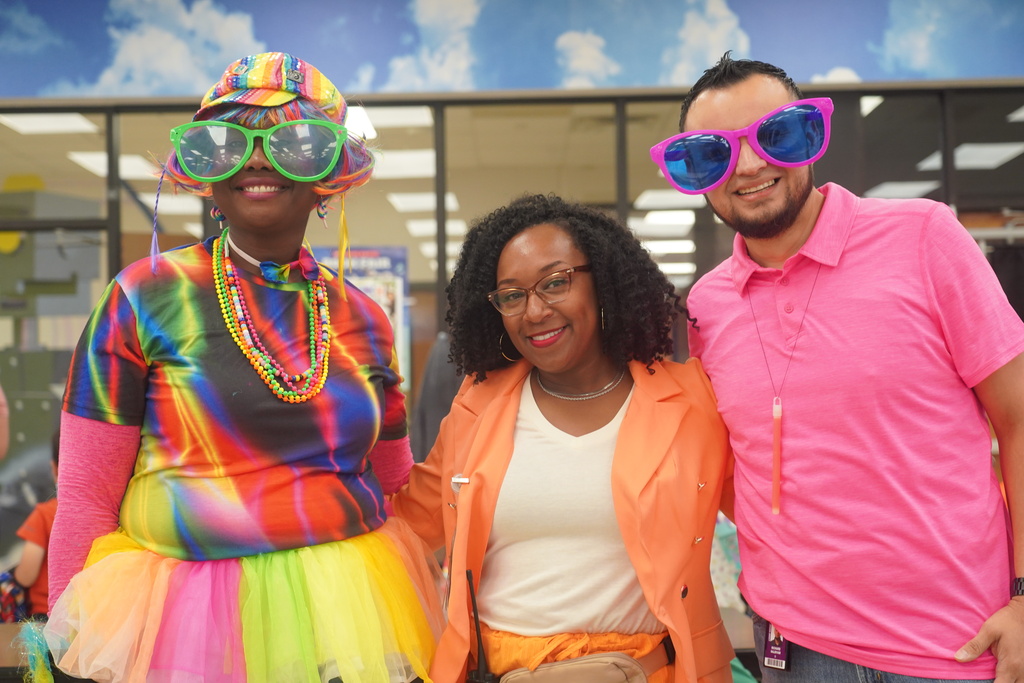 Three adults smile and pose for photo; one wears a tie dye dress, another an orange blazer.