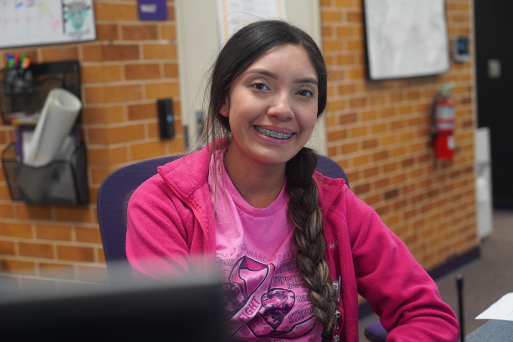 A student sits at a computer wearing a pink jacket and a pink shirt with a graphic.