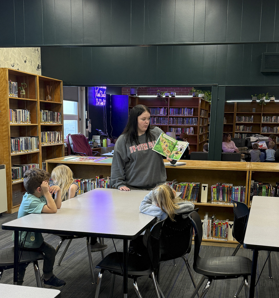 A woman reads a book to a group of children at a table in a library.