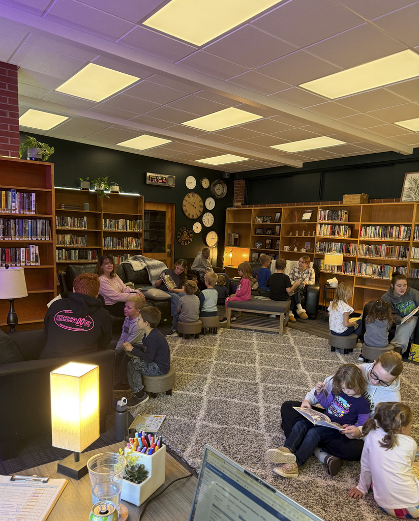 A library reading area with adults and children seated on stools, couches, and floor around tables.