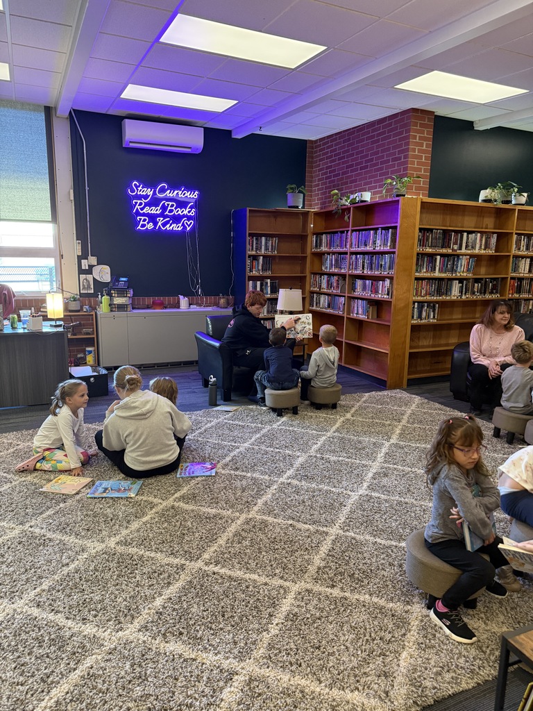 A library interior with several children sitting on a rug. A person reads aloud to the children.