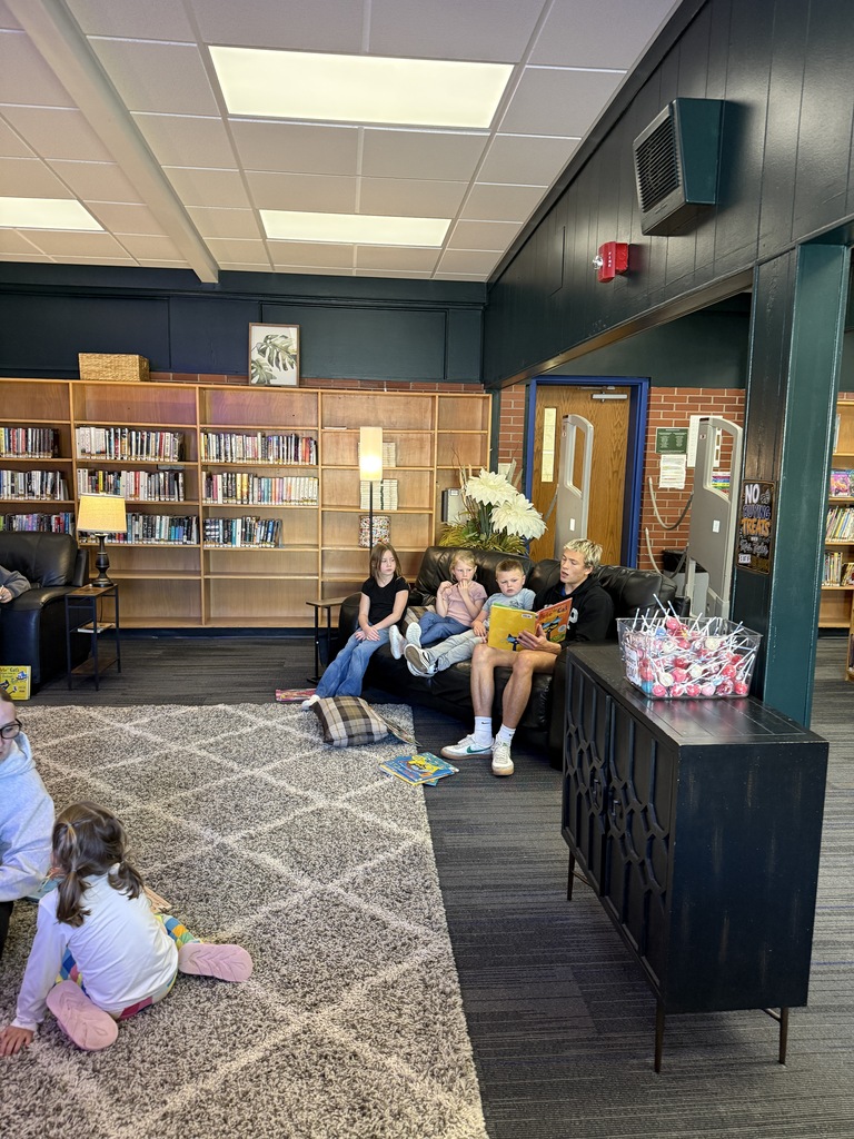 A group of people sit on a couch, with others sitting on the floor in a room with bookshelves.
