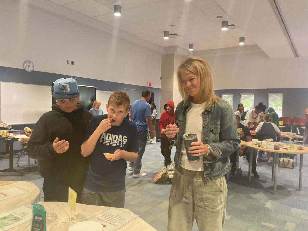 Two middle school boys sample something from a table in the Sampler room as a staff member stands by.