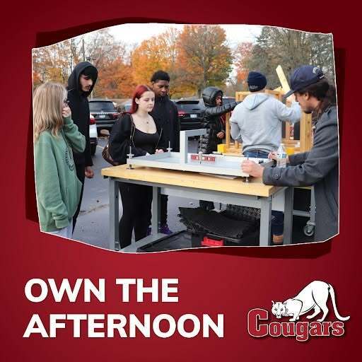 Group of students gathered around a table outdoors, engaging in a hands-on activity with autumn trees in the background.