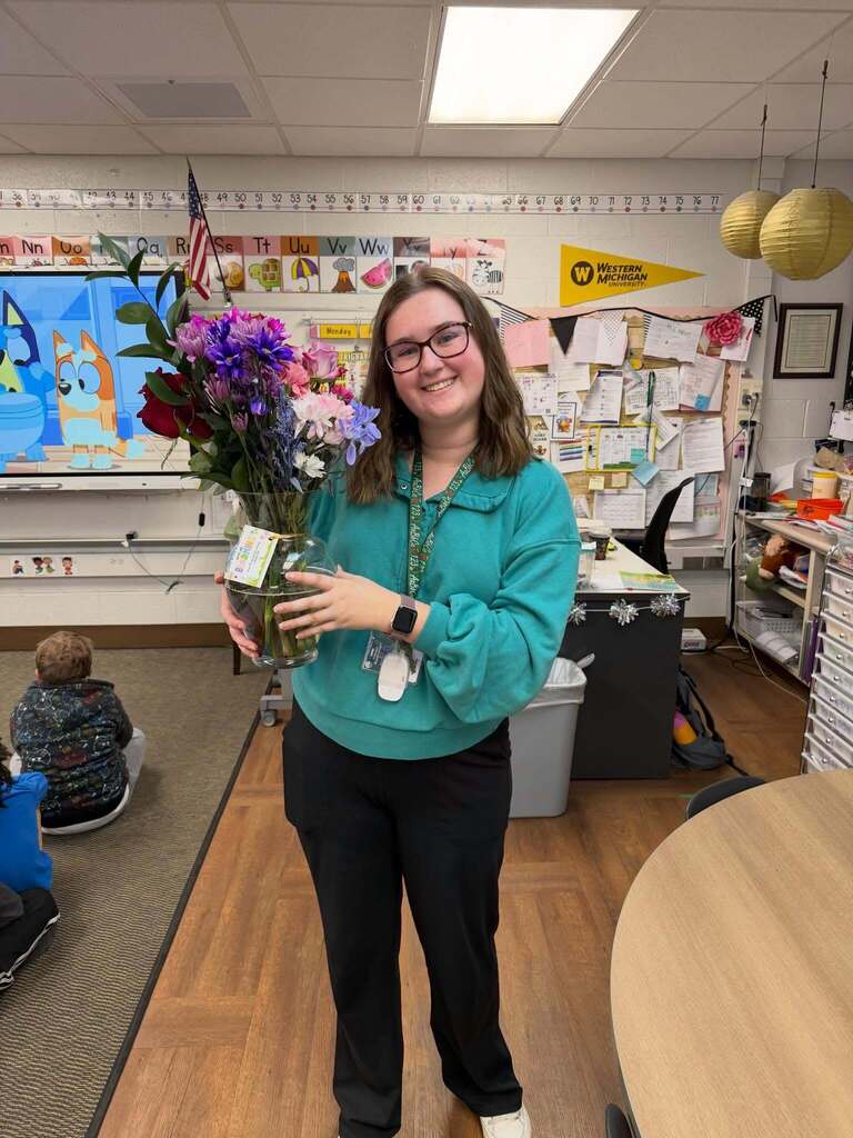 Teacher in a classroom holding a large bouquet of colorful flowers with educational posters and student work displayed behind her.