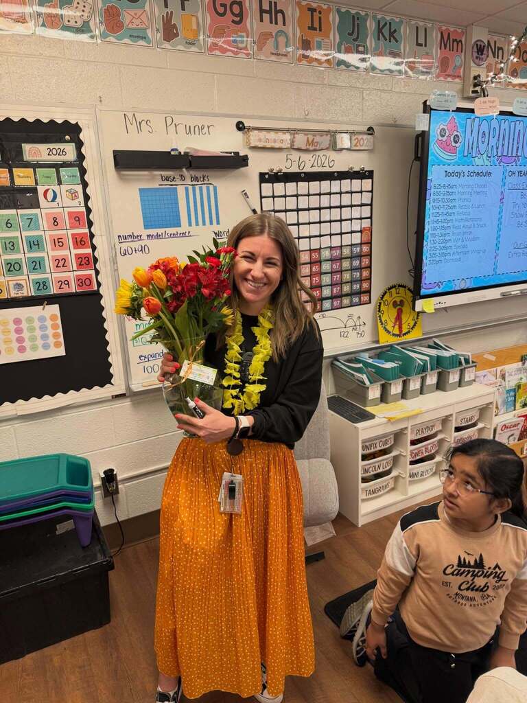 Teacher in a classroom holding a bouquet of flowers, wearing a yellow skirt and a lei, standing near educational charts and a digital board.