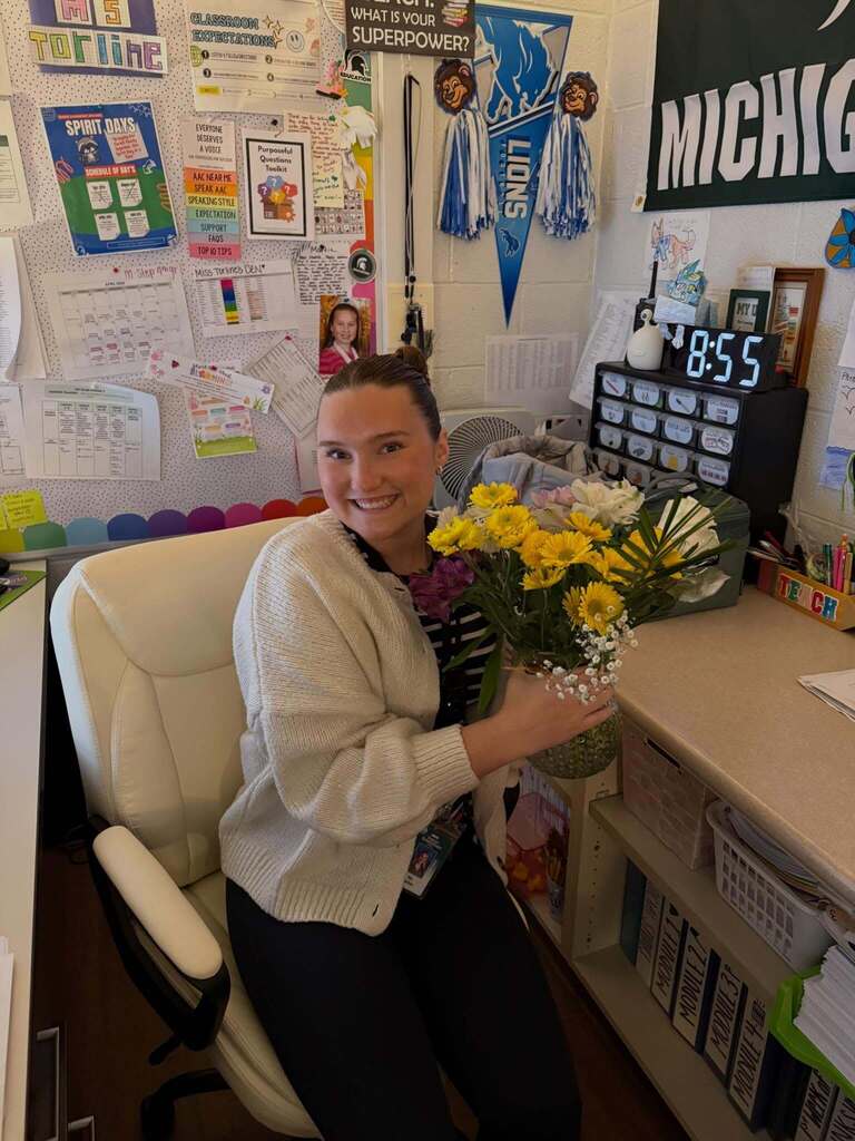 Person sitting in a classroom holding a colorful bouquet of flowers, surrounded by educational posters and a Michigan banner.