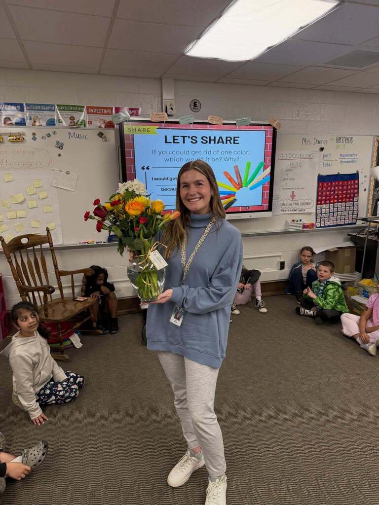 Teacher holding a bouquet of colorful flowers standing in a classroom with children seated on the floor around her.