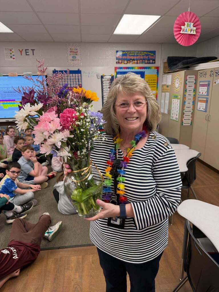 Teacher holding a large bouquet of colorful flowers in a classroom filled with seated young students.