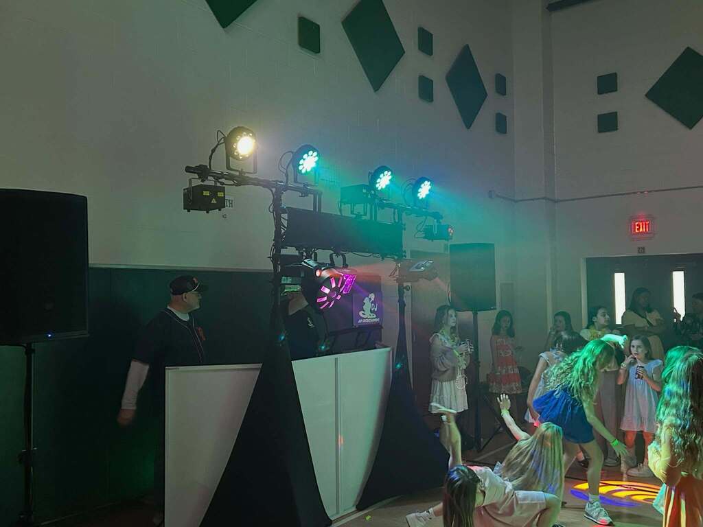 Children dancing and playing under colorful stage lights at an indoor event with a DJ setup in the background.