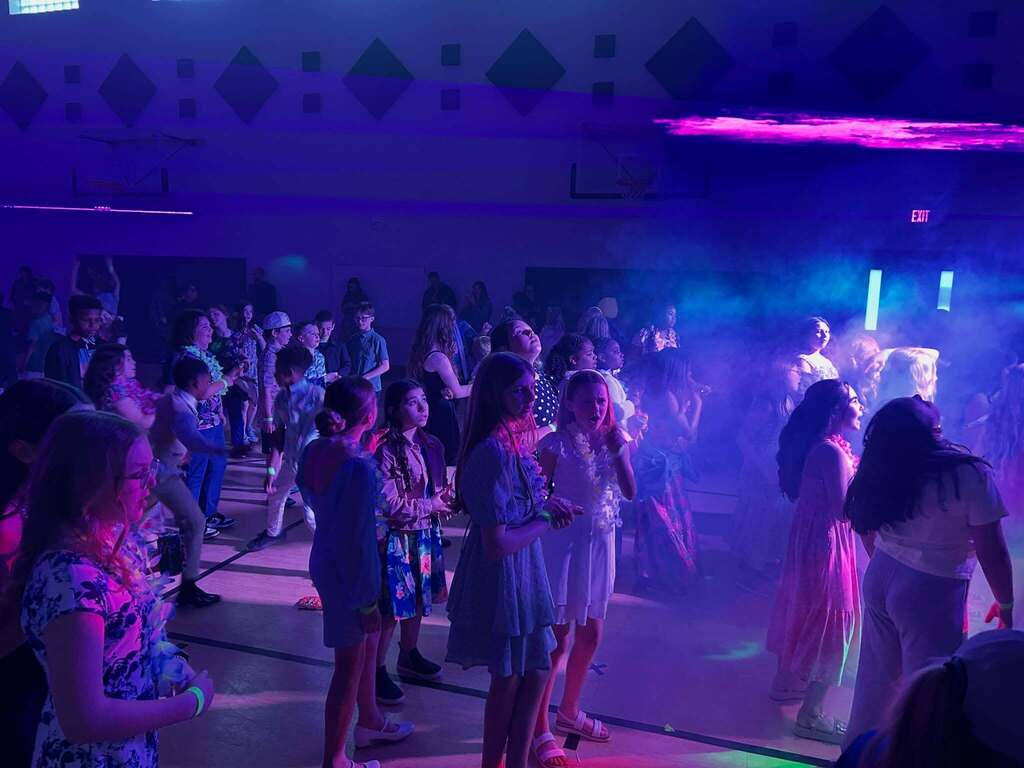 Group of young people dancing and socializing in a dimly lit room with colorful purple and blue lighting effects.