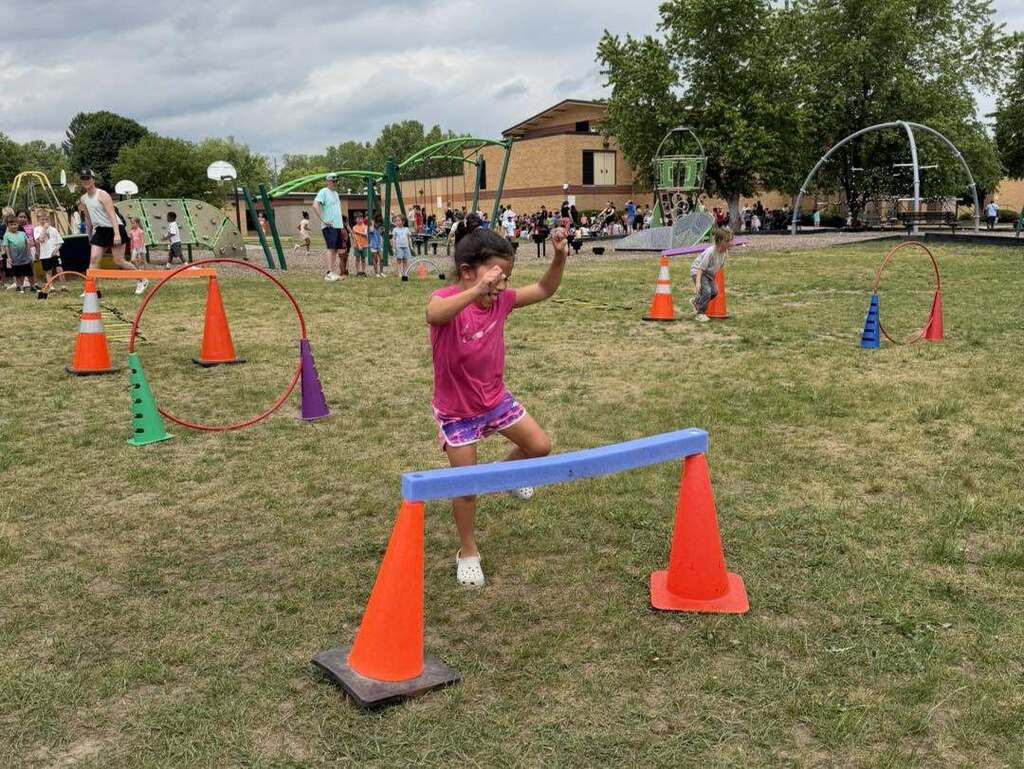 Child in pink shirt jumping over a blue bar supported by orange cones on a grassy field during an outdoor event.