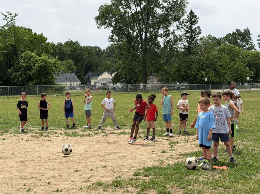Group of children standing on a grassy field preparing to play soccer with two balls visible on the ground.