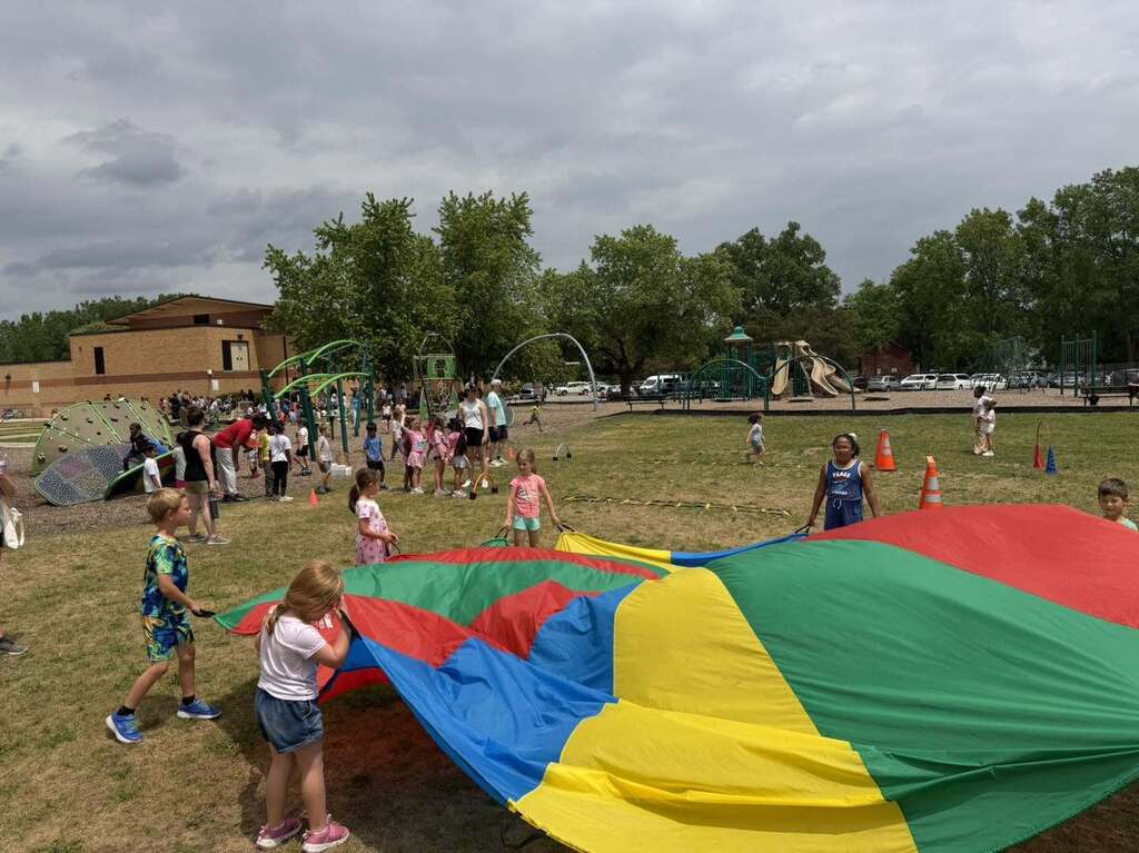 Children playing with a large colorful parachute on a grassy playground under a cloudy sky.