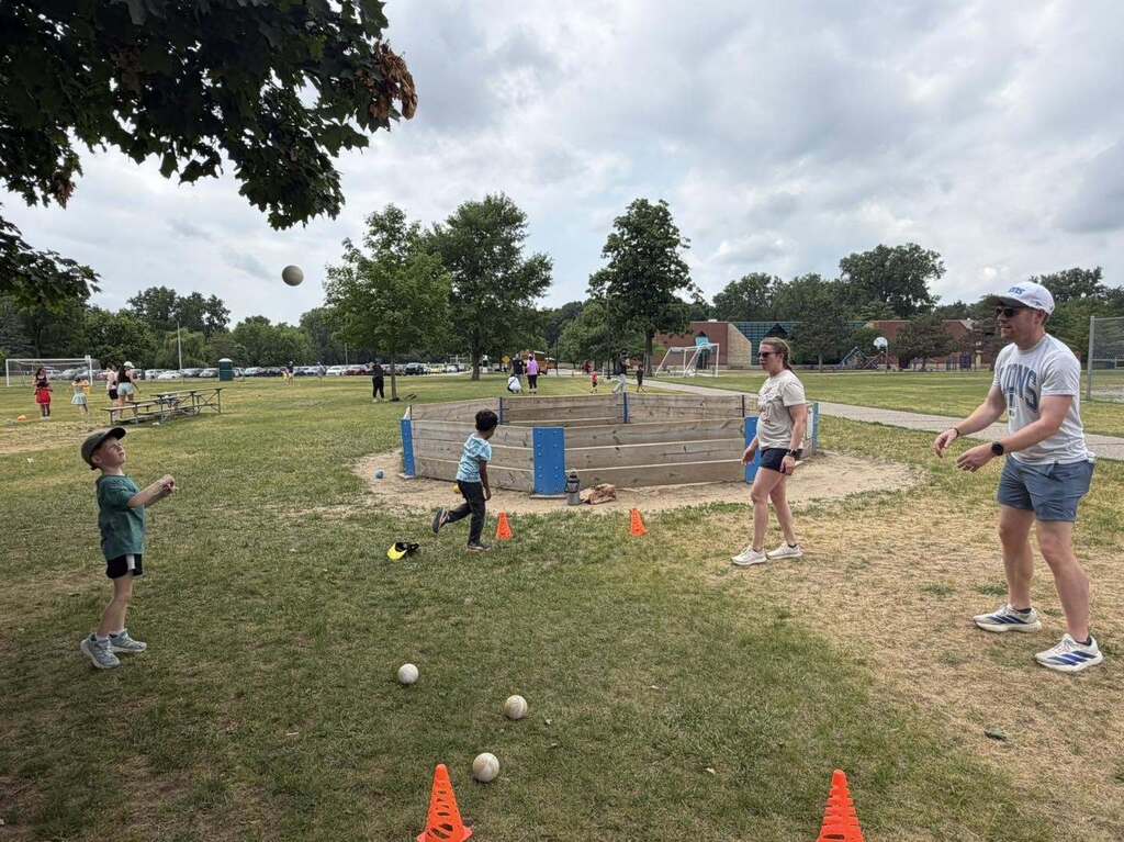 Children and adults playing a ball game on a grassy field with orange cones marking the play area under a cloudy sky.