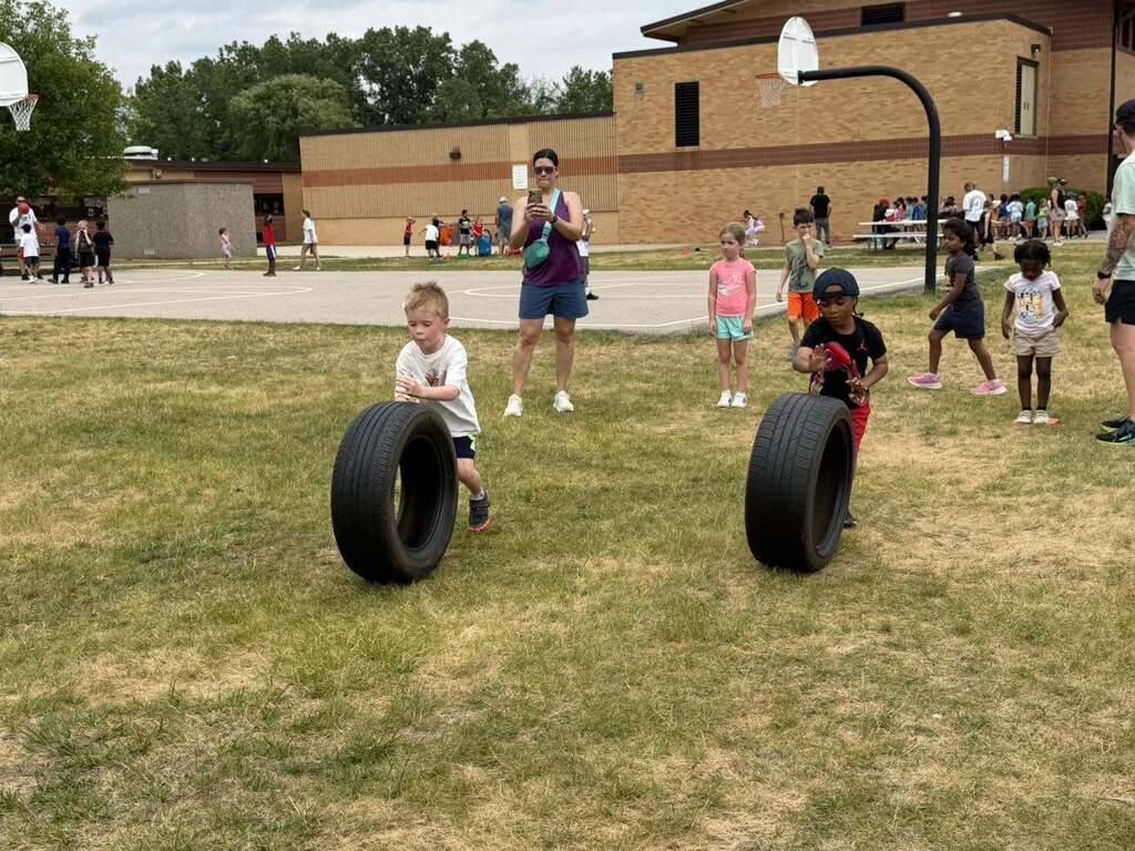 Two young boys race rolling large tires across a grassy field during an outdoor school event with other children and adults nearby.